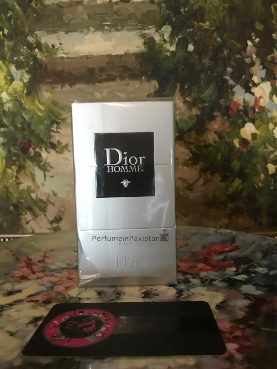 Dior Homme by Dior Eau de Toilette for men in white 100ml packaging displayed with PerfumeinPakistan branding card against an artistic floral background.
